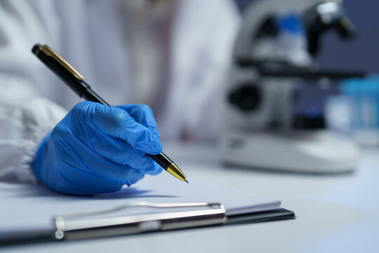 Close-up of scientist wearing blue gloves writing notes on clipboard in laboratory setting with microscope in background, emphasizing scientific research and documentation