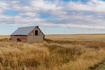 Abandoned barn in a field with wind turbines. Rural landscape under a cloudy sky. Empty, weathered barn stands amidst golden grasses. , Ririe, Idaho, USA.