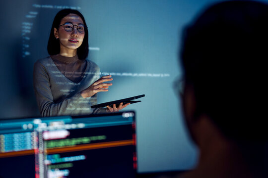 asian woman developers collaborating on a coding project, with one member presentation at code projected on the wall while the other works on multiple computer screens