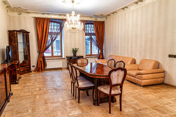 Well furnished living and dining room featuring a wooden dining table, beige leather sofa, chandelier, parquet flooring, and patterned curtains.