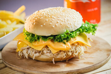 Photo of burger with sesame seed bun, chicken, salad and cheese surrounded by a soft drink and fries on a round wooden tray on the table of a wooden bar or restaurant next to a brick wall prepared for
