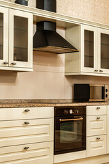 Modern kitchen featuring cream cabinets with glass doors, a black range hood, a built in oven with black and gold finish, and a speckled countertop.