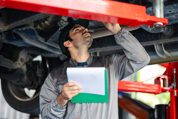 A skilled mechanic works under the vehicle and writes a detailed job checklist on a clipboard,...