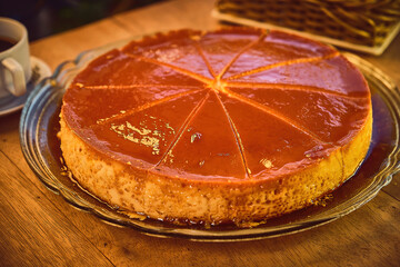 Photo of large, sliced ​​pudding in a glass tray on a wooden bar or restaurant table with a paper base and surrounded by a mug of coffee and a jar of sugar and a basket of condiments prepared for brea