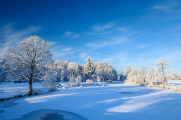 Winter landscape with frozen river surrounded by snow-covered trees illuminated by soft sunlight, casting shadows on snow. Eider river near Kluvensiek locks in winter, Rendsburg, Schleswig-Holstein.