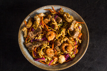 Yakisoba mixed with beef and shrimp, stuffed with assorted vegetables such as beetroot and carrots in a rustic plate on a black textured table-standard