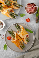 Baked puff pastry with green asparagus, prosciutto or ham and cheese on a gray concrete background. Top view.
