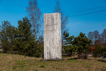 A weathered stone monument stands upright on grass, surrounded by trees with power lines in the background under a clear blue sky.