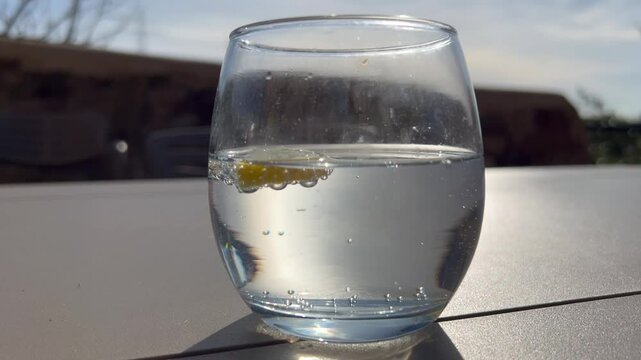 Video vaso de vidrio transparente relleno con t&oacute;nica y media luna de lim&oacute;n, la luz atraviesa el l&iacute;quido y las burbujas de gas se mueven. Fondo una mesa de jard&iacute;n y paisaje