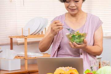 Senior Asian woman with short gray hair preparing healthy food in kitchen, using laptop listen to music or salad recipe research, modern lifestyle silver hair female in 70s friendly with technology