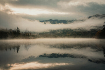 Fototapeta premium A calm lake surrounded by fog, with a faint reflection of trees and mountains in the still water, giving the scene a dreamy effect.