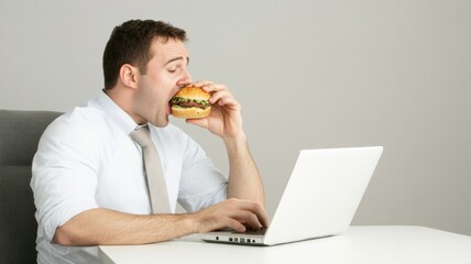 A man eating a burger while working on his laptop.