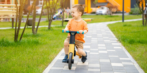 A cute happy toddler boy of two or three years old rides a bicycle or balance bike in a residential complex on a sunny summer day. Active kid playing outside. banner