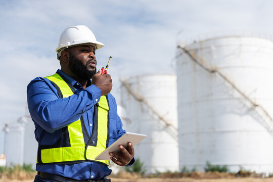 A safety engineer wearing a white hard hat, blue shirt, and yellow reflective vest uses a two-way radio while holding a tablet. The background features large industrial storage tanks under a blue sky