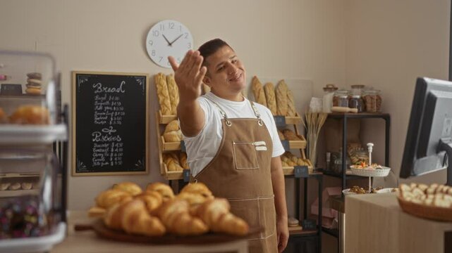 Young man in bakery wearing apron gesturing towards camera with hand, surrounded by various bread and pastries, clock in background, indoor setting, warm atmosphere