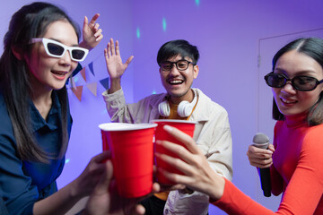 Fototapeta premium Group of happy Asian friends smiling at party, clinking glasses Young man and woman having fun at a house party, room lit with neon disco lights and dancing together, New Year's Eve party concept.