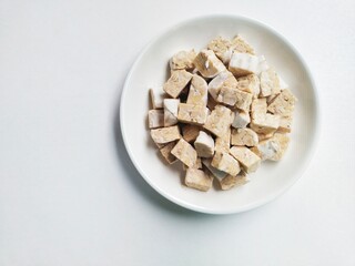 Uncooked tempeh pieces in a white plate on a white background. Top of view. 