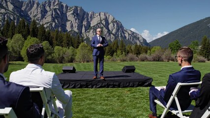 Outdoor ceremony featuring speaker in formal attire with stunning mountain backdrop in bright daylight