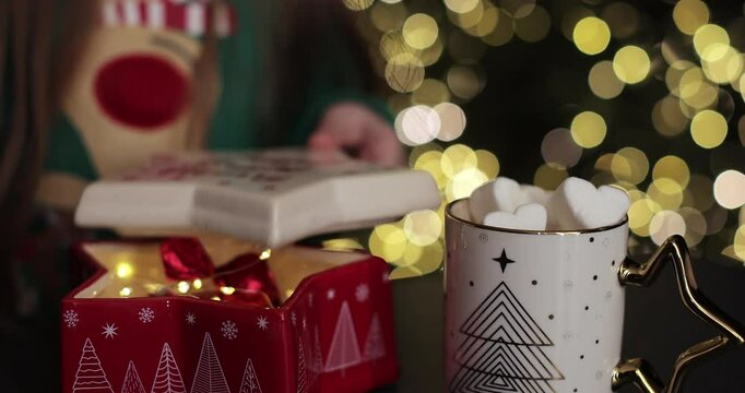 Little girl taking candies from Christmas jar and eating. Close-up hand. Decorated with lights and Christmas tree. Christmas mood.