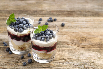 Tasty trifle dessert. Sponge cake, blueberries, jam and whipped cream in glasses on wooden table, closeup. Space for text