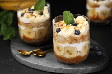 Tasty trifle dessert. Sponge cake, blueberries, banana and whipped cream in glasses on black table, closeup