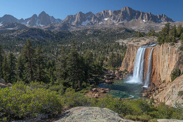 Waterfall cascades into a pool amidst a pine forest with a mountain backdrop.