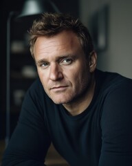 A serious Caucasian man in a black sweater poses thoughtfully in a softly lit room.