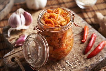 Fermented kim chi in a glass jar on a table with chilli peppers, sesame seeds, garlic and ginger