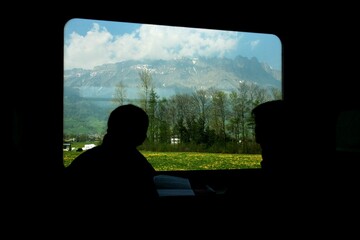 Silhouette of two people in a train with a scenic view of mountains and fields through the window.