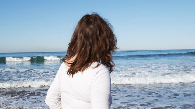 Mujer pelirroja disfrutando al caminar por la orilla de la playa, mira hacia el mar, surfista de fondo plano medio