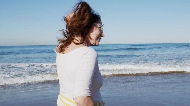 Mujer con gafas y pelo rojo, disfrutando al caminar por la orilla de la playa, se toca el pelo. Surfista de fondo plano corto