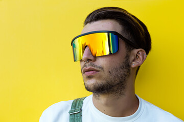 Isolated portrait of a young modern man wearing futuristic rainbow sunglasses, white t-shirt and a green overall in front of a yellow wall