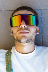 Isolated portrait of a young modern man wearing futuristic rainbow sunglasses, white t-shirt and a green overall lying on a concrete floor at the street