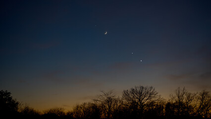 Crescent young Moon with stars, planets and countryside silhouettes.