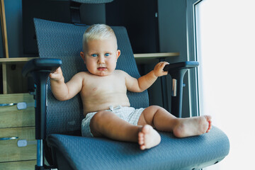 Toddler boy in shorts sits in a comfortable chair. The baby laughs and sits on a computer chair.