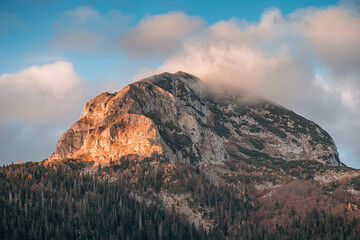 Golden hour sunlight illuminates a rugged mountain peak rising above a dense forest, with low...