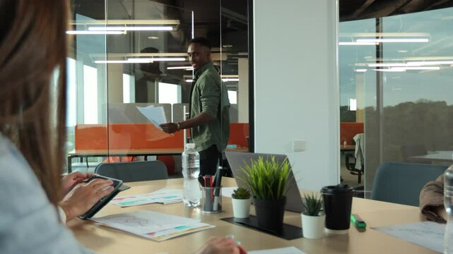 An african american hands out materials and schedules for discussion. Diverse startup company colleagues meeting in professional workplace, brifing and sharing ideas over financial strategy management