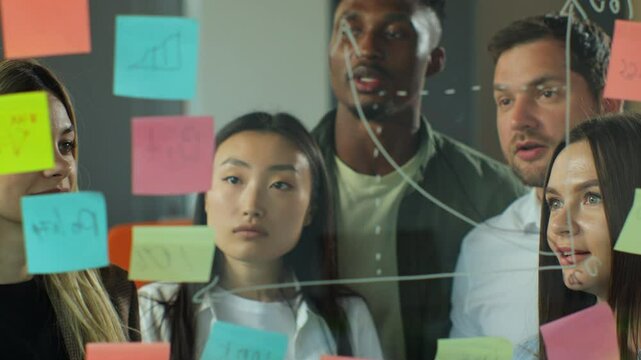 Diverse coworkers group analyzing corporate strategy drawing chart on glass wall with sticky notes. Multiracial business team people designers or marketing experts brainstorm at business meeting.