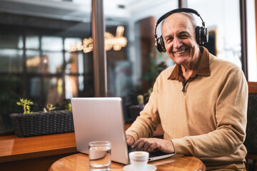 Fototapeta premium Smiling senior man in casual clothing with wireless headphones using laptop and drinking coffee while sitting in the cafe.