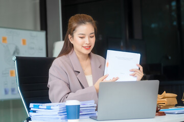 Attractive Asian businesswoman in formal business attire is working at her desk with a laptop and mobile phone, focusing on corporate law, contracts, compliance, and law firm marketing strategies.