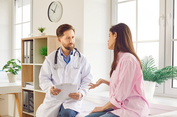 Fototapeta premium Portrait of a male doctor talking with young female patient sitting on couch in clinic in exam room. Physician giving consultation a woman during medical examination in office. Medicine concept.