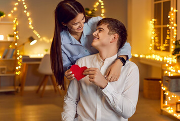 Happy couple looking at each other in the bedroom with garlands in the evening. Beautiful young woman giving a heart to a man. Concept valentine day, enjoy together, heart shape, romance