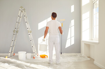 Young man painter from repair service in white clothes standing back in empty room and holding a...