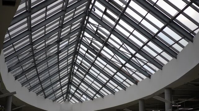 Spacious interior of a shopping mall featuring a large triangular glass ceiling supported by a metal framework. The design emphasizes modern and minimalist aesthetics. White walls and columns enhance