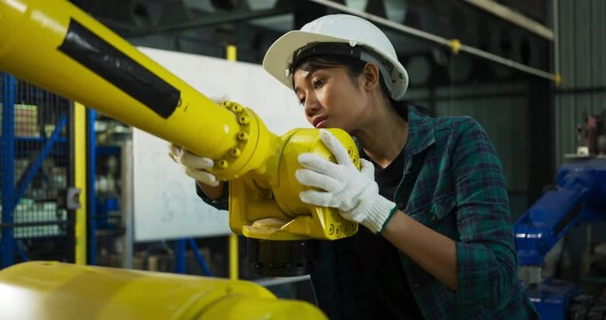 Focused female engineer wearing gloves and a hard hat, inspecting a yellow robotic arm in an industrial factory, showcasing technical expertise, precision, and dedication to robotics.