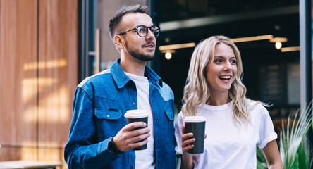 Happy caucasian male and female in trendy jeans denim wear standing together on street with coffee to go, positive hipster guys couple in love having fun and communicating during date in city