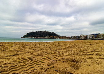 Sandy beach with waterfront buildings and hills in the background at sunset in San Sebastian, Spain. Coastal cityscape and natural landscape.