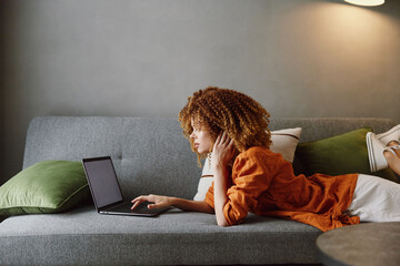Young woman with curly hair in an orange shirt working on a laptop on a cozy sofa, conveying focus and creativity with a relaxed ambiance in muted colors
