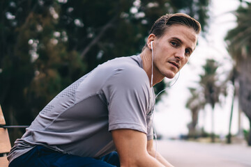Fototapeta premium Portrait of Caucasian male trainer sitting at urban bench and looking at camera whle listening audio songs from media app, athletic sportsman in digital earphones holding cellular and posing outdoors