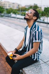 Satisfied young black hipster male with smartphone and takeaway coffee sitting on step in street